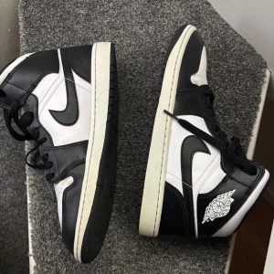 Jordan 1 mid 43 - Snygga Nike Air Jordans i klassisk svart och vit färgkombination. Skorna har Mid modell med snörning och den ikoniska Air Jordan-loggan på sidan. Perfekta för en stilren look.