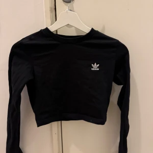 Svart croppad tröja från Adidas - Snygg svart croppad tröja från Adidas med deras ikoniska logga på bröstet. Tröjan har långa ärmar med de klassiska tre ränderna längs armen
