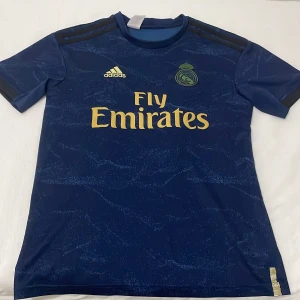 Blå Real Madrid fotbollströja från Adidas - Snygg blå Real Madrid fotbollströja från Adidas med Fly Emirates-logga i guld. Tröjan har korta ärmar och ett subtilt mönster över hela tyget. Perfekt för alla fotbollsfans!