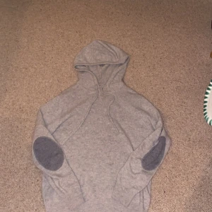 Grå hoodie i kashmir - Säljer en supermjuk och lyxig grå hoodie i 100% kashmir. Tröjan har en klassisk design med huva och mörkare armbågslappar som ger en snygg kontrast. Perfekt för kyliga dagar när du vill vara både bekväm och stilren.