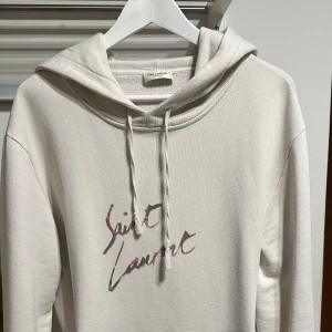 Beige hoodie från Saint Laurent - Ordinarie pris 7000kr. Snygg beige hoodie från Saint Laurent med deras ikoniska logga i en färgglad stil på bröstet. Tröjan har en klassisk känguruficka och justerbar huva med dragsko. 