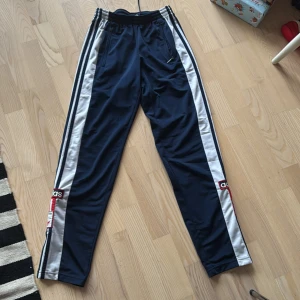 Blå träningsbyxor från Adidas - Snygga blå träningsbyxor från Adidas med vita och svarta ränder längs sidorna. Byxorna har knappar längs benen och en elastisk midja för extra komfort. Perfekta för en sportig look!