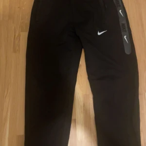 Svarta träningsbyxor från Nike - Snygga svarta träningsbyxor från Nike med vita logotyper längs benet. Byxorna har en bekväm passform och dragkedja vid fickorna för säker förvaring. Perfekta för träning eller en avslappnad dag.