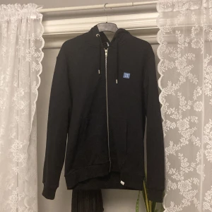 Svart hoodie från Les Deux - Snygg svart hoodie från Les Deux med dragkedja och en liten blå logga på bröstet. Perfekt för en casual look med sina långa ärmar och praktiska fickor framtill. Huvan har justerbara snören för extra komfort.