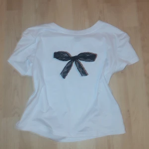 Vit t-shirt med svart rosett - Säljer en söt vit t-shirt med en svart rosett på ryggen. T-shirten har korta ärmar och en normal passform. Perfekt för en clean girl stil.