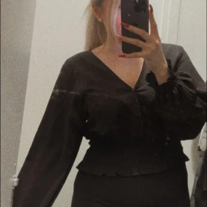 Svart omlottopp från Gina Tricot - Snygg svart omlottopp från Gina Tricot med långa ärmar och v-ringning. Toppen har en smockad midja och volangdetaljer vid nederkanten. Perfekt för en stilren look.