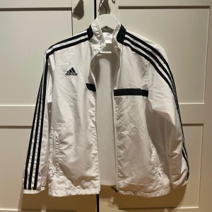 Vit vindjacka från Adidas - Säljer en stilren vit vindjacka från Adidas med svarta detaljer och de klassiska tre ränderna längs ärmarna. Jackan har dragkedja och två fickor framtill. Perfekt för sportiga aktiviteter eller en casual look. Ge gärna prisförslag 