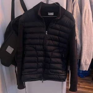 Moncler cardigan - Säljer en mörkblå cardigan från Moncler med dragkedja och långärmade detaljer. Jackan har en quiltad design och är perfekt för svalare väder. Den har en autenticitetskod för verifiering. Skicket är sådär samt ett hål i armen men det är därför jag säljer den så billigt.