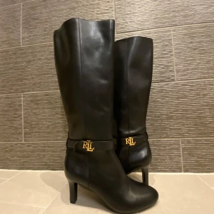 Svarta stövlar med klack från Ralph Lauren - Eleganta svarta stövlar i skinn från Ralph Lauren med klack och guldfärgad RLL-logga. Stövlarna har en dragkedja på insidan för enkel på- och avtagning. Stövlarna är i storlek 36/37 och helt oanvända då de är lite för små för mig. 