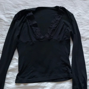 Svart långärmad topp med spetsdetaljer - Snygg svart långärmad topp med v-ringning och spetsdetaljer längs halsringningen. Perfekt för en stilren look.