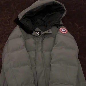 Grå Canada goose jacka  - Snygg grå dunjacka från Canada Goose.. Jackan har en quiltad design och stängs med dragkedja och knappar. Perfekt för kalla vinterdagar.                                                  Kan träffas i Stockholm eller frakta.
