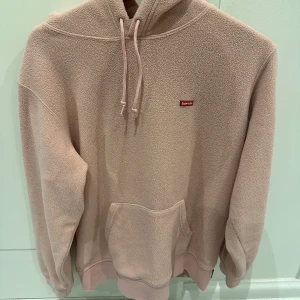 Rosa fleece hoodie från Supreme - Mysig rosa hoodie från Supreme i fleece-material. Den har en klassisk känguruficka och justerbar huva med dragsko. Perfekt för kyliga dagar.