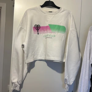 Vit croppad sweatshirt från Gina Tricot - Säljer en vit croppad sweatshirt från Gina Tricot med ett färgglatt tryck av tennisracketar och texten 'Court Club' på framsidan. Tröjan har långa ärmar och en avslappnad passform. 