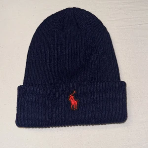 Mörkblå Ralph Lauren mössa - Helt oanvänd Snygg mörkblå stickad mössa från Polo Ralph Lauren med röd broderad logga framtill. Perfekt för kyliga dagar och ger en stilren look. Tillverkad i tjockt och mjukt material för extra värme och komfort.                                Priset kan diskuteras vid snabb affär!