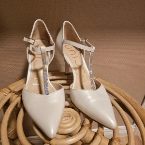 Vita klackskor från Unisa - Eleganta vita klackskor från Unisa med spetsig tå och glittrande rem över foten. Tillverkade i skinn med en stilren design, perfekta för speciella tillfällen. Använd endast vid två tillfällen. 