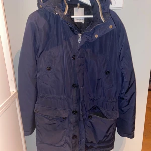 Mörk blå field jacket från J.Lindeberg - Snygg mörkblå field jacket från J.Lindeberg med huva och dragkedja. Jackan har knappar framtill och drag kedja över. Perfekt för kyligare dagar.