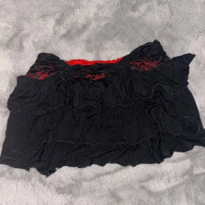 Svart kjol med spetsdetaljer - Trendig svart kortkjol med röda spetsdetaljer i midjan. Kjolen har volanger som ger en lekfull look. Perfekt för att sätta en unik touch på din outfit.