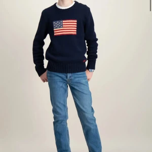 Mörkblå stickad tröja med amerikansk flagga - Snygg mörkblå stickad tröja med en stor amerikansk flagga på bröstet. Tröjan har långa ärmar och ribbade muddar vid ärmslut och nederkant. Perfekt för en .avslappnad stil.