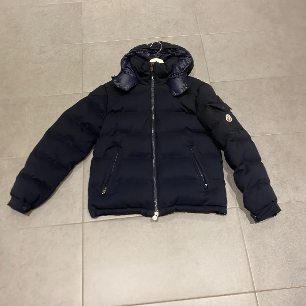 Säljer en snygg mörkblå dunjacka från Moncler med huva och dragkedja. Jackan har långa ärmar och praktiska fickor med knappar. Perfekt för kyliga dagar.. Takit.