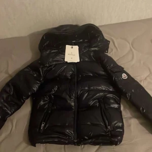 Moncler Maya Jacka - Säljer en snygg svart dunjacka från Moncler. Jackan har en glansig finish och är utrustad med en dragkedja och huva. Perfekt för kyliga dagar med sin varma och stilrena design. NFC funkar också 