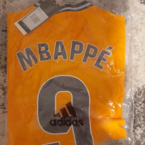 Real Madrid fotbollströja år 24/25 borta - Snygg orange Real Madrid-tröja från Adidas med Mbappé och nummer 9 på ryggen. Tröjan har korta ärmar och klubbens emblem på bröstet tillsammans med sponsorloggan 'Emirates Fly Better'. Perfekt för fotbollsfans! ST M passar dig runt 175-185cm om du är runt 75-85KG