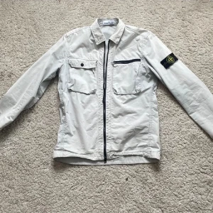 BYTE  - Snygg vit overshirt från Stone Island med dragkedja och två bröstfickor. Den har en klassisk krage och en broderad logga på ärmen. Perfekt för lager-på-lager-stil. Pris kan diskuteras vid riktigt snabb affär 