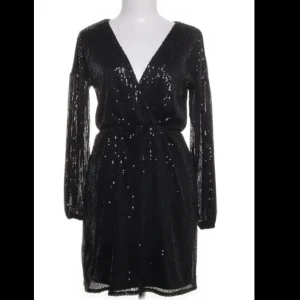 Svart glittrig klänning från Dennis Maglic - Snygg svart klänning från Dennis Maglic med glittriga paljetter och omlott-design. Klänningen har långa ärmar och ett knytband i midjan för en smickrande passform. Perfekt för en festlig kväll!
