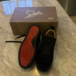 Svarta sneakers från Christian Louboutin - Snygga svarta sneakers från Christian Louboutin med den ikoniska röda sulan. Skorna har snörning och är tillverkade i mocka, vilket ger en lyxig känsla. Perfekta för den som vill ha en stilren look med en touch av exklusivitet. Enden på snöret är sönder på två. Sulan ör lite sliten på grund av mycket användning.