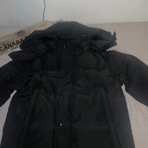 Svart dunjacka från Canada Goose - Säljer en svart dunjacka från Canada Goose med avtagbar pälskrage. Jackan har en klassisk design med lång ärm och knappar. Perfekt för kalla vinterdagar.Den är helt ny aldrig använd den har kvar påse o allt med sig. Vill bli av med den för den passar inte mig
