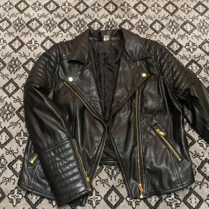 Svart skinnjacka från H&M Divided - Snygg svart skinnjacka från H&M Divided med dragkedjor och quiltade detaljer på ärmarna. Jackan har en klassisk bikerstil med guldfärgade dragkedjor och tryckknappar vid kragen.