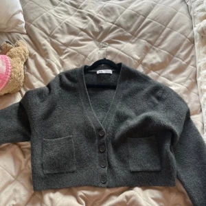 Grå kofta från Zara - Säljer en stilren grå kofta från Zara med knappar framtill och två praktiska fickor. Koftan har en v-ringad design och är perfekt för kyligare dagar. Den är croppad och har en mjuk och bekväm känsla.