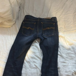 Mörkblå jeans från Nudie Jeans - Säljer ett par mörkblå jeans från Nudie Jeans i ekologisk bomull. De har en klassisk femficksdesign med snygga sömmar på bakfickorna. Perfekta för dig som gillar en stilren look med en touch av hållbarhet.