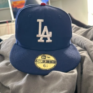 Blå keps från New Era - Snygg blå keps från New Era med LA-logga i vitt framtill. Perfekt för att komplettera din streetwear-look. Kepsen har en rak skärm och är i storlek 6 7/8.