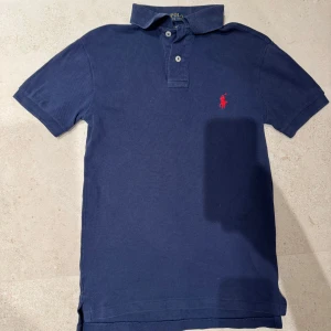 Mörkblå pikétröja från Ralph Lauren - Säljer en stilren mörkblå pikétröja från Ralph Lauren i slim fit. Tröjan har korta ärmar och en klassisk krage med knappar. Den ikoniska röda loggan är broderad på bröstet. Perfekt för en avslappnad men ändå snygg look. Skriv vid minsta fundering!