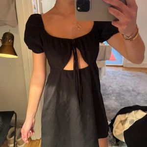 Svart klänning med knytband - Trendig svart klänning med korta puffärmar och knytband framtill. Den har en liten öppning vid bysten som ger en unik detalj.