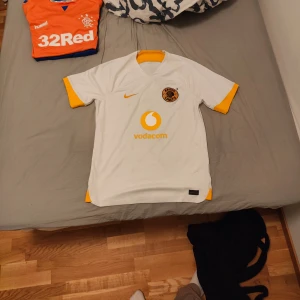 Kaizer Chiefs bortatröja 22/23 - Snygg vit fotbollströja från Nike med gula detaljer på ärmar och krage. Tröjan har en logga på bröstet och är kortärmad, perfekt för match eller träning. Den har även en sponsormärkning på framsidan. Skick: 8/10