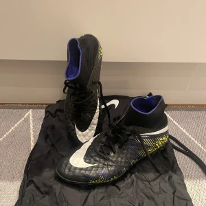Nike Hypervenom Elite SG - Snygga fotbollsskor från Nike i svart med gula, blåa och vita detaljer. Skorna har en hög ankelkrage och snörning för bra passform. Sulan har ett iögonfallande gult mönster med dobbar för optimalt grepp på planen. Inga hål eller separationer.