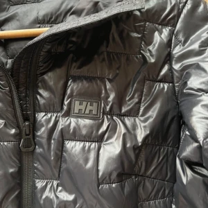 Svart pufferjacka från Helly Hansen nypris 2300 kr - Säljer en svart pufferjacka från Helly Hansen med dragkedja framtill. Jackan har ett quiltat mönster och är perfekt för kyligare väder. Den är långärmad och har en stilren design med HH-logga på bröstet.