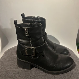 Svarta boots med dragkedja och bälte - Snygga svarta boots i skinn med dragkedja och dekorativt bälte. Perfekta för höst och vinter med en robust klack för extra komfort.  Från skopunkten