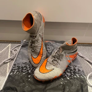 Nike Hypervenom FG - Säljer ett par grå och orange fotbollsskor från Nike med hög ankel och snörning. Skorna har en iögonfallande design med svarta detaljer och en mönstrad sula för bra grepp på planen. Snörena har släppts längst ner på höger fot och även ett litet hål längst fram på skon (se sistabild). Snörena har fixats så funktionen påverkas ej. OBS! Inga innersulor finns.
