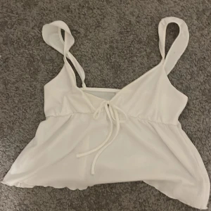 Vit ärmlös topp med knytband - Säljer en söt vit ärmlös topp med knytband framtill. Toppen har en omlott-design och breda axelband, perfekt för varma dagar.
