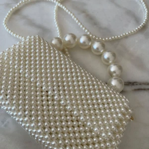 Vit pärlväska - Elegant kuvertväska helt täckt av vita pärlor med ett handtag av större pärlor. Perfekt för att ge en lyxig touch till din outfit. Väskan har en kompakt design och är idealisk för speciella tillfällen.
