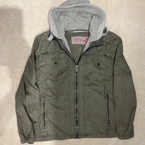 Grön Overshirt med Huva - Snygg grön jacka med grå huva och dragkedja. Jackan har flera fickor med knappar och ribbade ärmslut. Perfekt för en casual look. Vid minsta fråga eller fundering hör av er🤝