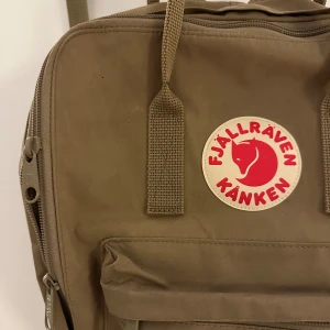 Beige ryggsäck, Fjällräven Kånken - Säljer en klassisk beige/brun Fjällräven Kånken ryggsäck. Den har justerbara axelremmar, ett stort huvudfack och en framficka med dragkedja. Perfekt för vardagsbruk och utflykter. Ikonisk logga på framsidan.