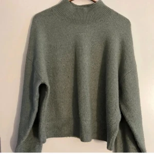 Stickad tröja i alpackaull  - Mysig grön stickad tröja från & Other Stories med hög krage och långa ärmar. Perfekt för kyliga dagar med sin avslappnade passform och ribbade detaljer vid krage, ärmslut och nederkant.