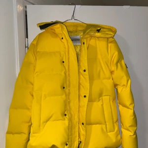 Gul dunjacka från Kenzo - Snygg gul dunjacka från Kenzo med huva och dragkedja. Jackan har långa ärmar och en stilren design med Kenzo-logga på insidan och ärmen. Perfekt för att lysa upp vinterdagarna!