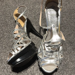 Silvriga klackskor med remmar - Snygga silvriga klackskor med remmar och öppen tå från  Bianco . Skorna har en hög klack och en platåsula i svart. Perfekta för en festlig kväll.