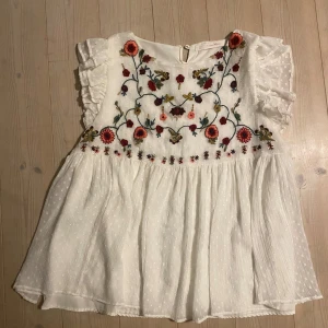 Vit blus med broderade blommor från Zara - Charmig vit blus från Zara med vackra broderade blommor i flera färger. Blusen har volangdetaljer vid ärmarna och en knappstängning bak. Perfekt för en romantisk look.
