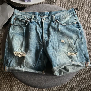 Blå jeansshorts från Levi's - Snygga blå jeansshorts från Levi's med slitna detaljer och uppvikta ben. Perfekta för en avslappnad stil. Klassisk femficksdesign och knappgylf. Storlek W31