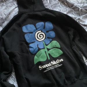 Svart hoodie från Sweet SKTBS - Säljer en svart hoodie från Sweet Sktsb med en stor, färgglad blomma på ryggen i blått och grönt. Hoodien har en klassisk design med ficka och snör-luva. Perfekt för en avslappnad stil! Har ALDRIG används!!! Är i storlek M så passa både killar o tjejer ☺️